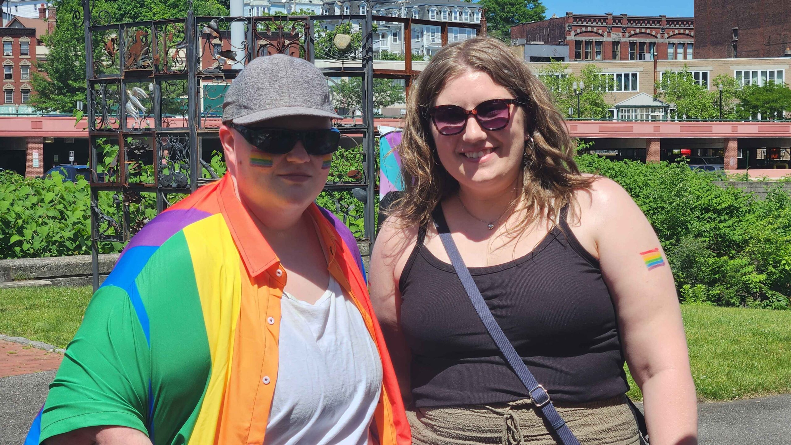 Two People standing side by side. Person on the left is wearing a collared shirt like a rainbow pride flag and a gray hat with sunglasses. Person on the right is wearing a black tank top and sunglasses.